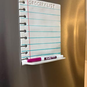 May include: A white ceramic magnetic dry-erase board shaped like a piece of notebook paper with the text GROCERY LIST embossed at the top. It features horizontal teal lines, a vertical red margin line, spiral binding holes on the left, and a small shelf holding a magenta Expo dry-erase marker.