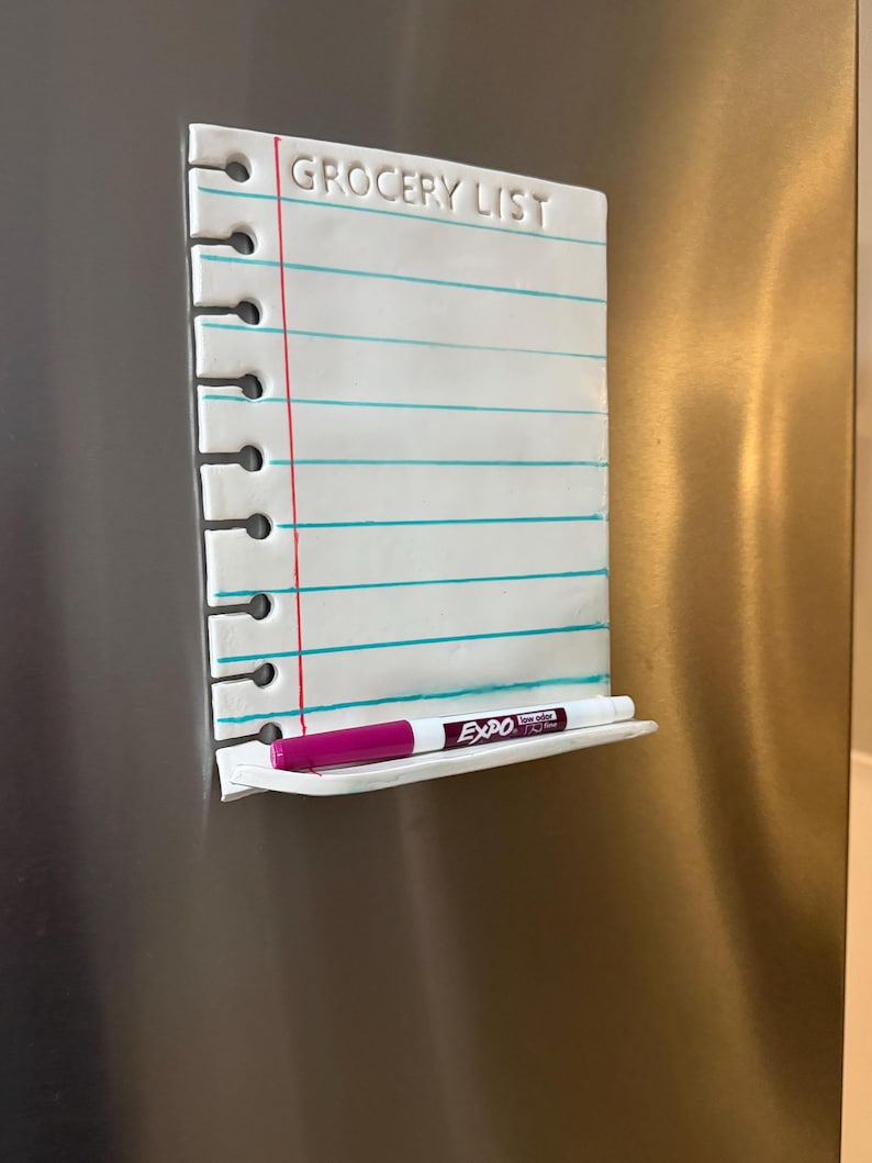 May include: A white ceramic magnetic dry-erase board shaped like a piece of notebook paper with the text GROCERY LIST embossed at the top. It features horizontal teal lines, a vertical red margin line, spiral binding holes on the left, and a small shelf holding a magenta Expo dry-erase marker.