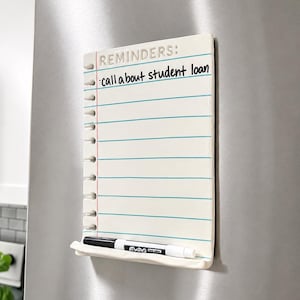 May include: A white ceramic magnetic dry-erase board shaped like a piece of notebook paper with blue horizontal lines and a red vertical margin line. The text REMINDERS: and call about student loan are written on it. A black dry-erase marker rests below.