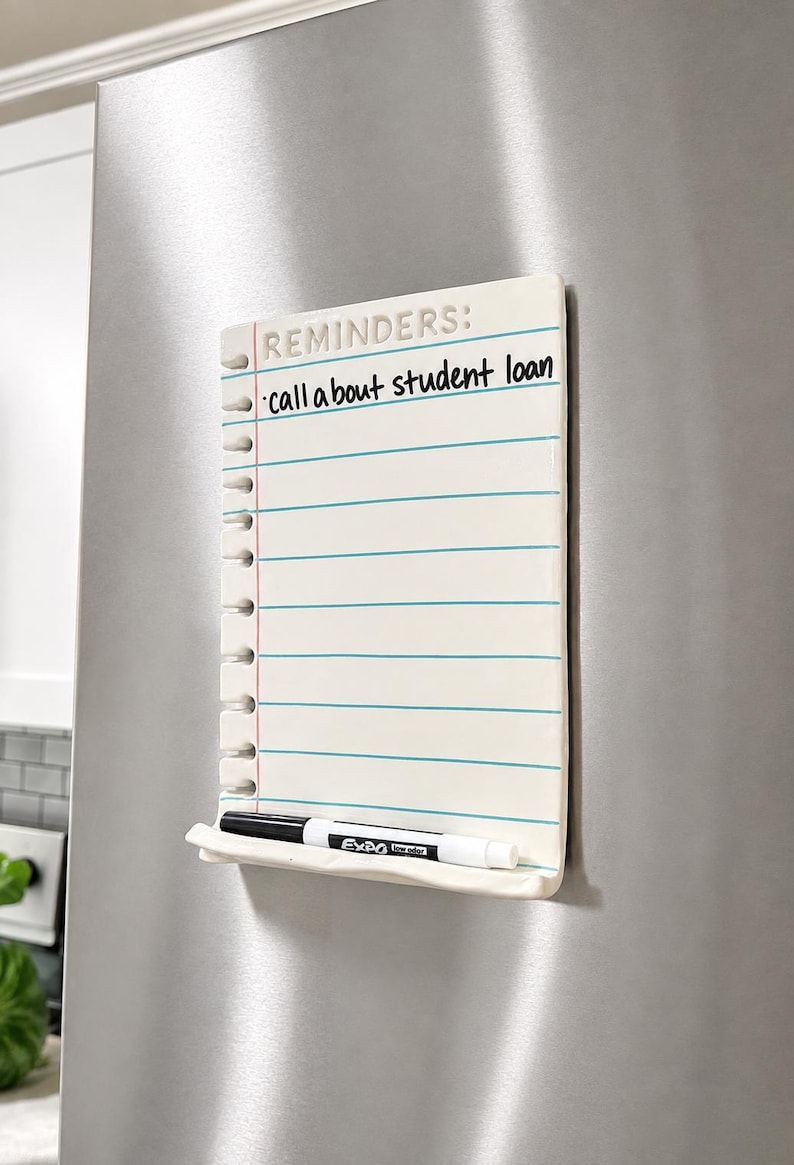 May include: A white ceramic magnetic dry-erase board shaped like a piece of notebook paper with blue horizontal lines and a red vertical margin line. The text REMINDERS: and call about student loan are written on it. A black dry-erase marker rests below.