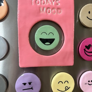 May include: A rectangular pink magnet frame with the text TODAYS MOOD embossed at the top and a circular cutout in the center. A light green circular magnet with a black laughing face emoji is positioned inside the frame, surrounded by other colorful circular magnets with various facial expressions.