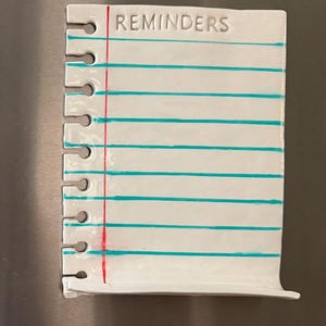 May include: A white ceramic notepad magnet shaped like a piece of spiral-bound notebook paper. It features the word REMINDERS at the top, a vertical red margin line, and several horizontal teal lines. The left side has cutouts for spiral binding.