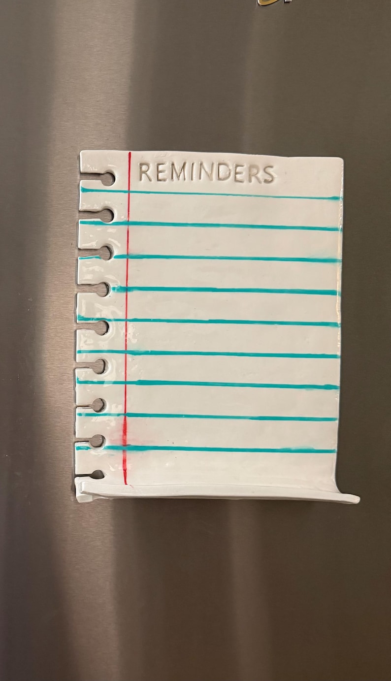 May include: A white ceramic notepad magnet shaped like a piece of spiral-bound notebook paper. It features the word REMINDERS at the top, a vertical red margin line, and several horizontal teal lines. The left side has cutouts for spiral binding.