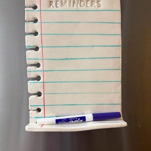 May include: A white ceramic dry erase board shaped like a piece of notebook paper with the word REMINDERS embossed at the top. It features horizontal teal lines, a vertical red margin line, a spiral-bound edge, and a blue Expo dry erase marker.