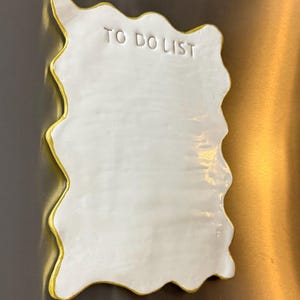 May include: A white ceramic magnetic memo board with an irregular, wavy edge and a gold-painted border. The surface features the embossed text TO DO LIST at the top, providing a blank space for notes on a smooth, glossy, and reflective finish.