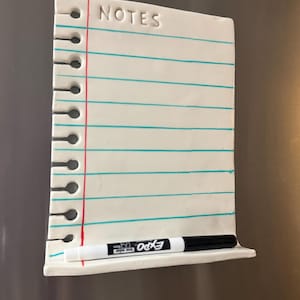 May include: A white ceramic magnetic notepad shaped like a piece of notebook paper with the word NOTES embossed at the top. It features horizontal teal lines, a vertical red margin line, and a small shelf at the bottom holding a black dry-erase marker.