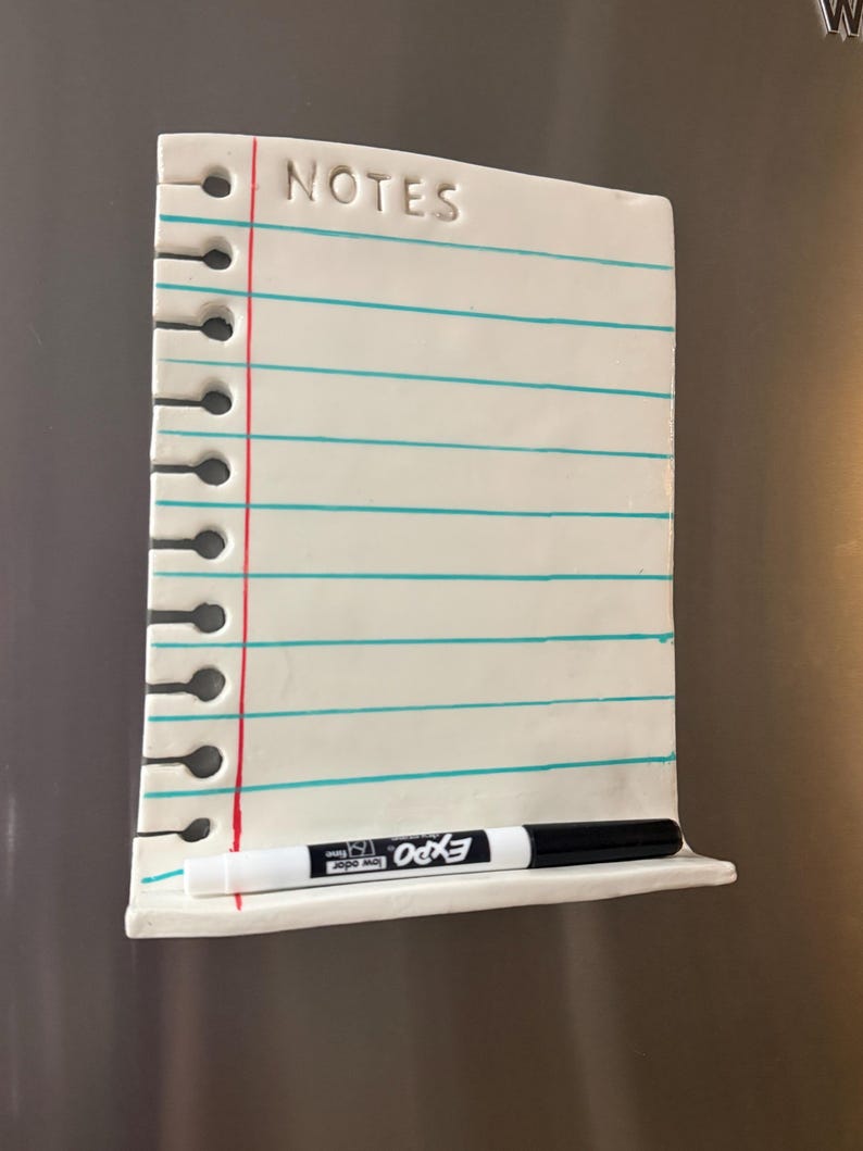 May include: A white ceramic magnetic notepad shaped like a piece of notebook paper with the word NOTES embossed at the top. It features horizontal teal lines, a vertical red margin line, and a small shelf at the bottom holding a black dry-erase marker.