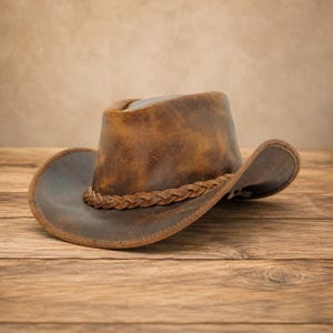 May include: A brown leather cowboy hat with a distressed finish and a braided leather band encircling the base of the crown. The hat features a wide, curved brim and a structured, rounded crown, showcasing natural variations in the leather texture.