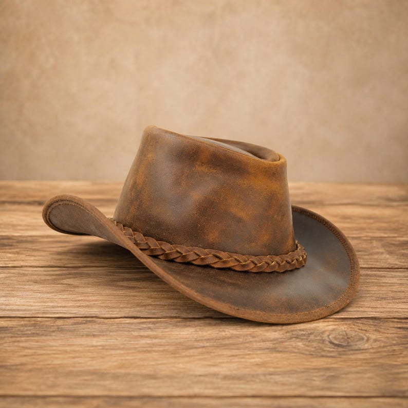 May include: A brown leather cowboy hat with a distressed finish and a braided leather hatband. The hat features a high crown and a wide, curved brim, showcasing natural variations in the leather texture and color throughout the surface.
