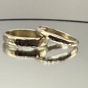 May include: Two gold-toned rings with a hammered, textured surface finish. The rings are positioned side-by-side, with one band being noticeably wider than the other. Both rings feature a reflective, metallic sheen and a rounded profile.