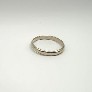 May include: A polished, solid gold-toned metal band ring with a smooth, rounded surface and a continuous circular shape. The ring features a reflective, metallic finish that catches the light, showcasing a simple, unadorned design.