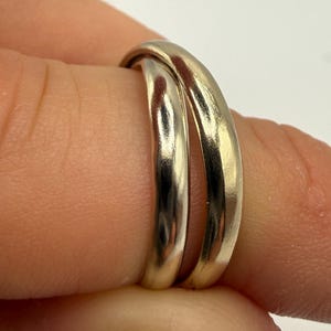May include: A pair of interlocking metal rings with a smooth, polished finish. The rings feature a rounded, domed profile and a reflective surface that catches the light, showing a consistent metallic tone across both bands.