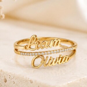 Può includere: Un anello color oro con due fasce orizzontali collegate da una fila centrale di piccole pietre trasparenti. La fascia superiore riporta il nome Liam in corsivo, mentre la fascia inferiore riporta il nome Olivia in corsivo coordinato.