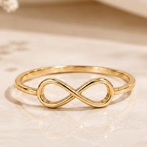 Peut inclure: Une bague en or poli présentant un motif central en forme de symbole de l'infini. L'anneau est fin et circulaire, se connectant aux boucles symétriques du motif. Le métal possède une surface lisse et réfléchissante qui capte la lumière.