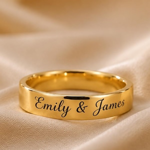 Può includere: Un anello a fascia in tonalità oro lucido con il testo inciso Emily & James in un carattere corsivo. L'anello presenta una superficie liscia e riflettente e un profilo piatto, mostrando un design pulito con scritte nere chiare e leggibili.