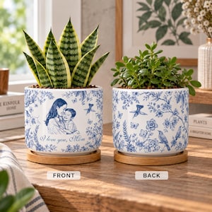 May include: Two white ceramic cylindrical planters with blue floral patterns and wooden saucers. The left planter features a sketch of a person holding a child with the text I love you, Mom. The right planter displays birds and flowers.