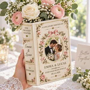 May include: A cream-colored ceramic book-shaped vase featuring a central oval photo of a couple, floral accents, and text including "Our Greatest Chapter," "Emma & Lucas," "1st Anniversary," "A Love Story Written By," and "Every Page Begins With Us 08.14.2026."