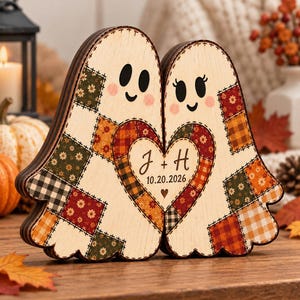 May include: A wooden decorative piece featuring two ghost shapes joined together, decorated with a patchwork pattern of autumn-colored squares. A central heart contains the text J + H and the date 10.20.2026 with a small heart icon below.