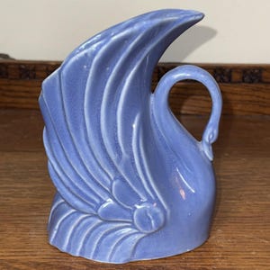 Puede incluir: Un jarrón de cerámica azul bígaro con forma de cisne, acabado brillante y detalles de alas moldeadas. El cuello del cisne forma un asa curva, mientras que el cuerpo del ave sirve como recipiente, mostrando patrones de plumas en relieve.