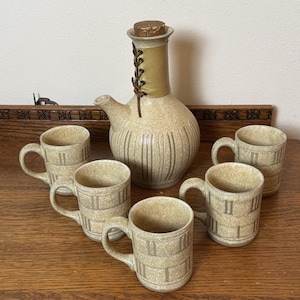May include: A ceramic beverage set consisting of a round decanter with a cork stopper and a tan leather lace-up neck detail, accompanied by five matching mugs. Each piece features a speckled beige glaze with vertical and horizontal line patterns.