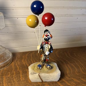 May include: A clown figurine standing on a rectangular stone base, holding three glossy balloons in blue, yellow, and red. The clown wears a black jacket, a white shirt with a red polka-dot bow tie, and metallic gold trousers with blue shoes.