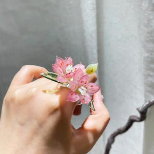 May include: A ring featuring two translucent pink flowers with delicate red speckles and white centres. The floral design includes green leaves and a small yellow bud, all crafted with a glossy finish that catches the light, creating a vibrant look.