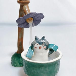 May include: A ceramic incense burner featuring a brown post with a purple flower top that releases smoke. A small, grey and white cat-like figure with a happy expression sits inside a textured green basin with three small brown feet.