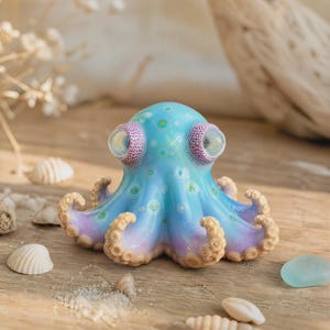 Può includere: Una piccola statuetta di polpo con una finitura liscia e lucida, caratterizzata da una sfumatura dal blu chiaro al viola tenue. La testa è decorata con piccoli e intricati motivi circolari bianchi e gialli e presenta due grandi occhi rotondi e chiari.
