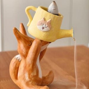 以下が含まれることがあります： 頭の上に黄色いジョウロを掲げた陶器製のキツネの置物。ジョウロには猫の顔のイラストが描かれており、中には火のついたお香のコーンが入っている。注ぎ口からは細い煙が流れ出し、台座の方へと向かっている。