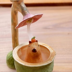 Puede incluir: Un quemador de incienso de cerámica con un capibara marrón claro sentado dentro de un cuenco verde texturizado. Una pequeña naranja descansa sobre la cabeza del capibara. Arriba, una pieza rosa en forma de flor sostiene un cono de incienso.