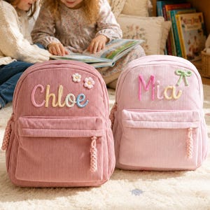 May include: Two corduroy backpacks in dusty rose and light pink. The dusty rose backpack features the name Chloe in embroidered letters with two small flower patches. The light pink backpack features the name Mia with a small green bow patch.