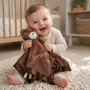 May include: A brown plush security blanket featuring a teddy bear head with a cream-coloured muzzle. The soft fabric is embroidered with the name George and a paw print, finished with a dark brown satin border and small fabric ribbon tags.