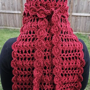 May include: A burgundy crochet head covering and shawl. The head covering is a fitted cap with an openwork design. The shawl features a scalloped edge and a decorative floral pattern down the back. The handmade item is worn over a black top.