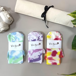 Könnte beinhalten: Drei Paar gerippte Socken mit Batikmustern in Blau und Grün, Lila und Weiß sowie Gelb und Pink. Jedes Paar ist mit einer weißen Papierbanderole befestigt, die den schwarzen Text Viva Barre und ein kleines Blumensymbol zeigt.