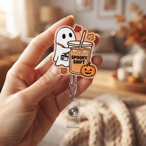 May include: An acrylic badge reel featuring a white ghost character holding an iced coffee cup with the text SPOOKY SHIFT printed on it. The design includes a small orange jack-o'-lantern and scattered autumn leaves on a glittery peach background.