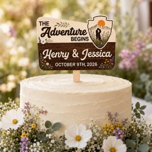 May include: A wooden cake topper featuring the text "THE Adventure BEGINS" and "Henry & Jessica" with the date "OCTOBER 9TH, 2026". The design includes a mountain landscape graphic with a sun, a couple silhouette, and floral accents.