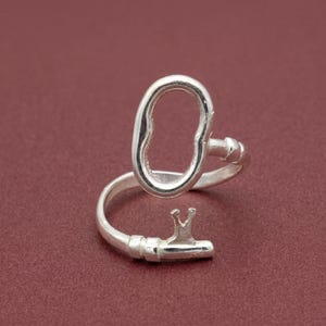 Pode incluir: Um anel aberto de tom prateado com um design de chave estilizado. O anel tem um acabamento liso e polido com uma cabeça circular e uma pequena extremidade dentada que imita o segredo de uma chave, criando uma estrutura de banda ajustável.