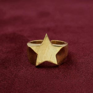 May include: A gold-toned ring featuring a prominent five-pointed star design on the front. The star has a brushed metal texture, while the band of the ring is smooth and solid, creating a cohesive piece of jewelry with a metallic finish.