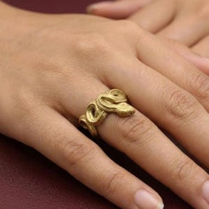 Può includere: Un anello color oro a forma di serpente avvolto, caratterizzato da una superficie testurizzata che imita le squame. Il serpente si avvolge attorno al dito, con la testa posizionata lateralmente e la coda che si curva per chiudere l'anello.