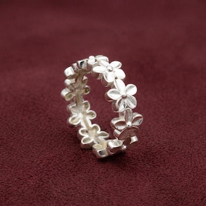 Può includere: Un anello in argento caratterizzato da una fascia continua di piccoli fiori a cinque petali collegati tra loro. Il metallo ha una finitura lucida e il motivo floreale si ripete lungo l'intera circonferenza della fascia, creando un aspetto delicato.