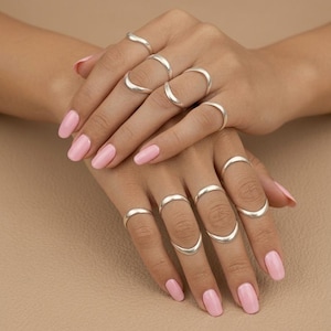 Può includere: Una collezione di dieci anelli in metallo color argento liscio indossati sulle dita. Ogni anello presenta un design semplice a fascia curva con una finitura lucida. Gli anelli sono distribuiti su entrambe le mani, alcuni posizionati sulle falangi.