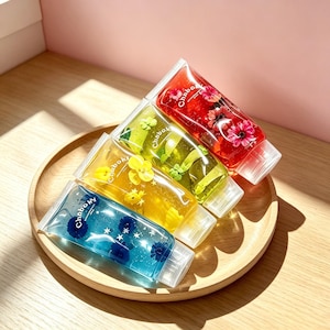 Può includere: Quattro tubetti trasparenti riempiti di gel colorato, ognuno con elementi floreali incorporati e piccole forme a stella. I tubetti riportano la scritta Chnboky e sono disposti in una pila diagonale, mostrando tonalità rosse, verdi, gialle e blu.