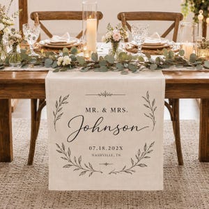 May include: A beige linen table runner featuring black printed text that reads MR. & MRS. Johnson, the date 07.18.202X, and the location Nashville, TN. The design is framed by two symmetrical botanical leaf branches in a circular wreath style.