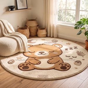 Op de afbeelding: Een rond, crèmekleurig vloerkleed met een grote centrale illustratie van een slapende bruine beer. Het ontwerp bevat kleine sterren, een halve maan en de tekst "TheClassicsRugs" in kleine letters gedrukt bij de kop van de beer.