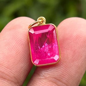 May include: A rectangular pendant featuring a vibrant hot pink gemstone set in a polished gold-toned metal frame. The pendant includes a small circular gold-toned bail at the top for attachment to a chain, showcasing a faceted surface design.