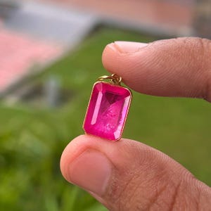 Op de afbeelding: Een rechthoekige hanger met een levendige, doorschijnende felroze edelsteen in een eenvoudige goudkleurige metalen zetting. De hanger is voorzien van een klein rond goudkleurig oogje aan de bovenkant voor bevestiging aan een ketting en heeft een facetgeslepen afwerking.