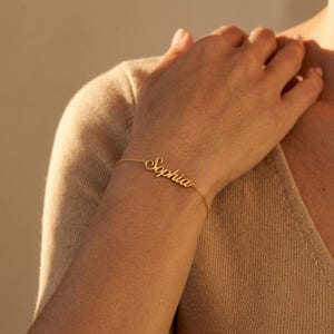 Op de afbeelding: Een goudkleurige kettingarmband met een centraal naamplaatje waarop de naam Sophia in een cursief lettertype staat. De armband bestaat uit een fijne schakelketting die om de pols sluit en een gepolijste metalen afwerking heeft.