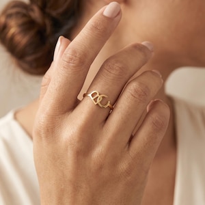 Könnte beinhalten: Ein goldfarbener Ring mit zwei ineinandergreifenden Großbuchstaben, B und C, als zentrales Designelement. Das Band ist dünn und poliert und zeigt eine minimalistische Ästhetik, wobei die Buchstaben nebeneinander auf dem Finger liegen.