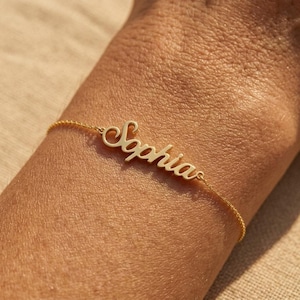 May include: A gold-colored metal bracelet featuring a delicate chain and a central nameplate with the name Sophia written in a cursive script font. The piece is designed with a polished finish and connects to a thin chain on both sides of the name.