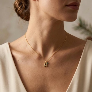May include: A gold-toned necklace featuring a delicate chain with a pendant consisting of a capital letter A charm and a small, round, dark green gemstone charm. The necklace rests against the collarbone, showcasing a polished metallic finish.